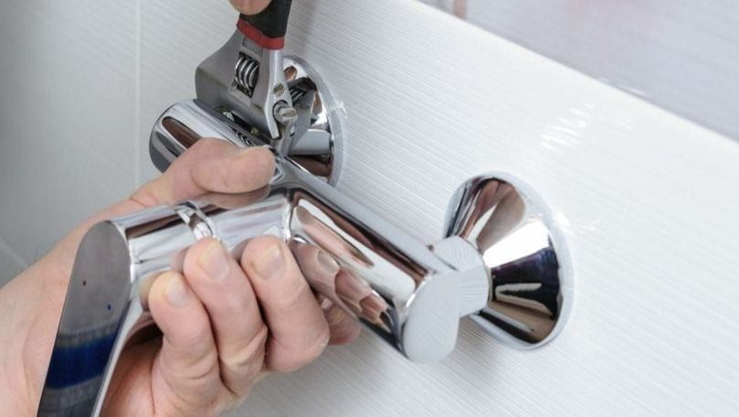 Person uses a wrench and screwdriver to install a shiny chrome faucet/handle into a wall flange (close-up).