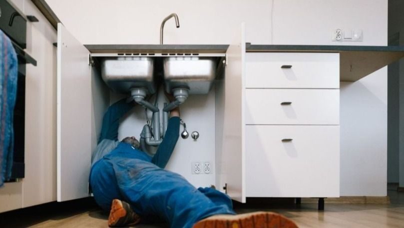 A plumber lying on the floor under a double kitchen sink, working on exposed pipes beneath white cabinetry.