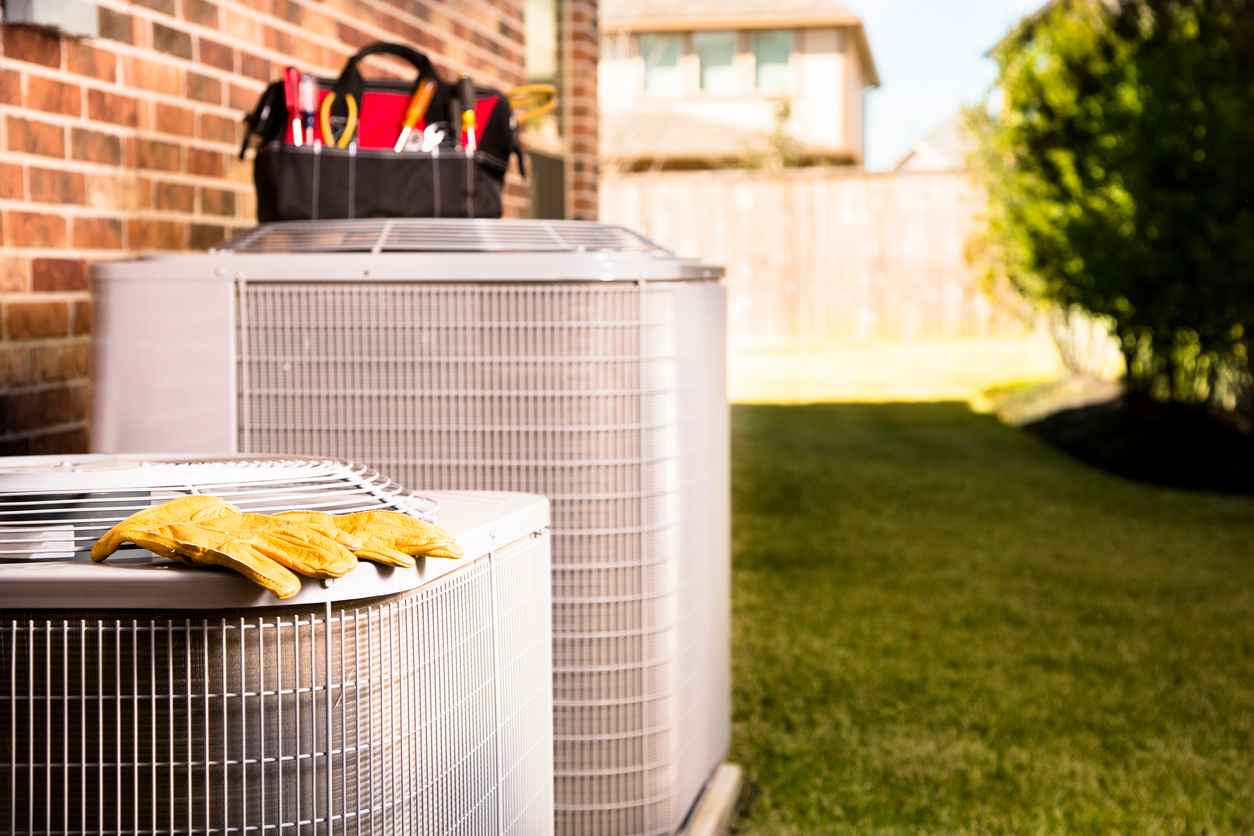 Bag of repairman's work tools, gloves on top of air conditioner units outside a brick home. Service industry, working class.