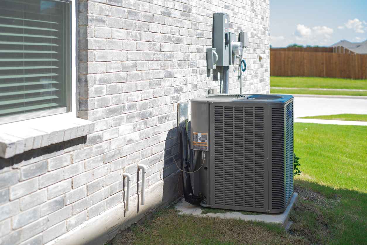 Side windows of new construction home and central air conditioning condenser unit mounted on concrete slab beside brick house wall