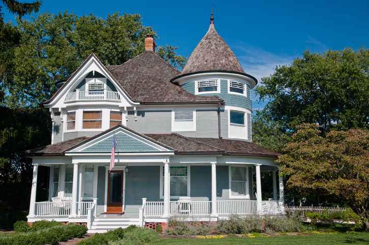 Beautiful gray traditional victorian house. House has an American Flag hanging over the porch and shows a beautiful garden with flowers and trees.