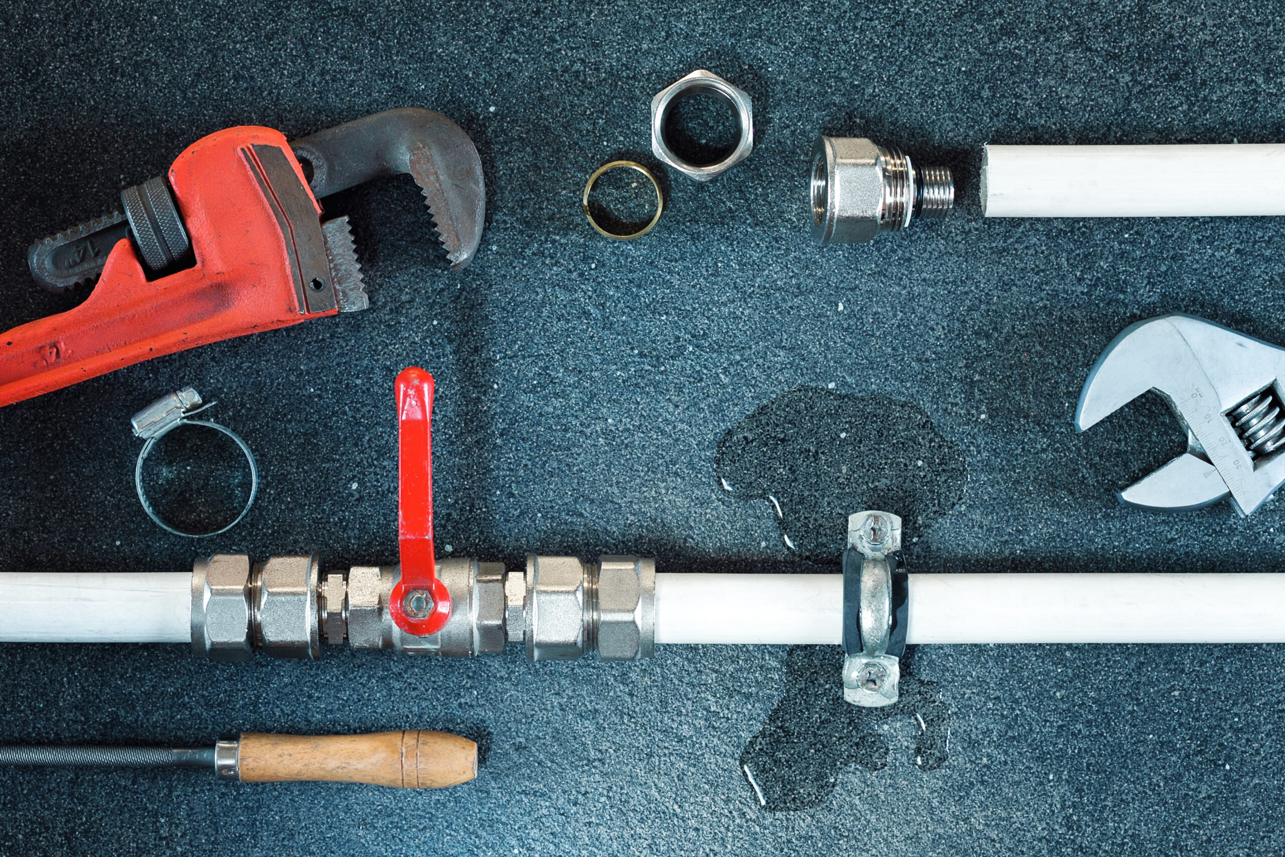 Assortment of plumbing tools and fittings on a dark surface, including a red pipe wrench, nuts, and PVC pipe pieces.