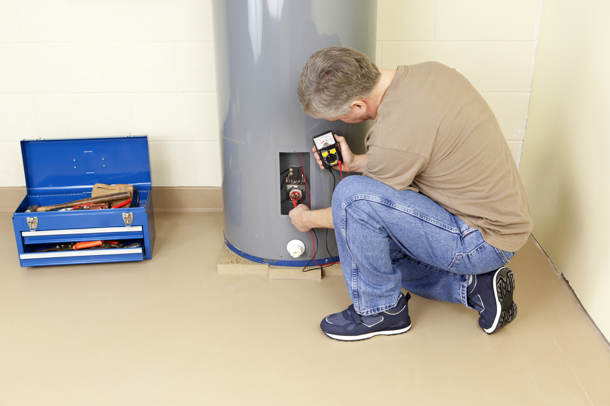 Technician kneels by a tall gray water heater, testing electrical connections with a digital multimeter; a blue toolbox lies open nearby.
