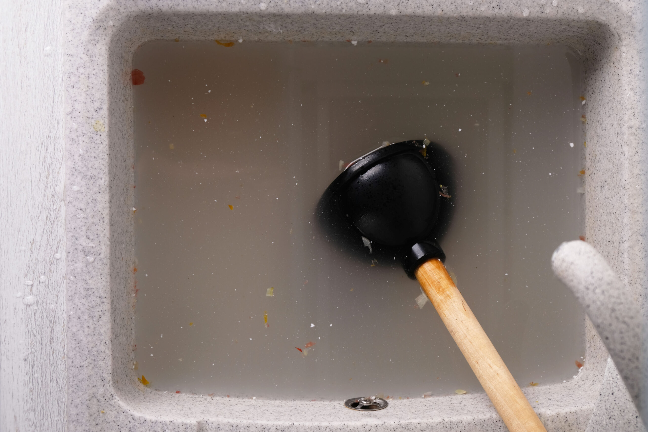 Top-down view of a gray kitchen sink filled with muddy water and a black rubber plunger with a wooden handle inside.