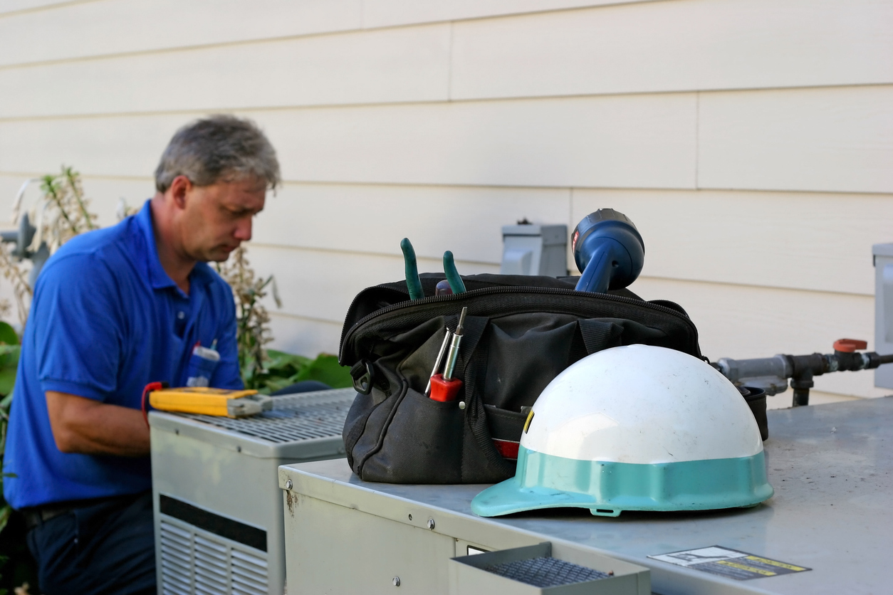 HVAC technician working on outdoor AC unit. His hat and toolkit are sitting on top of the unit.