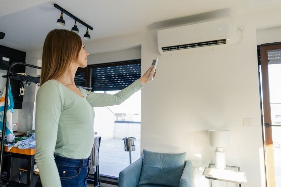 Woman in a mint sweater points a remote at a wall-mounted air conditioner in a bright living room.