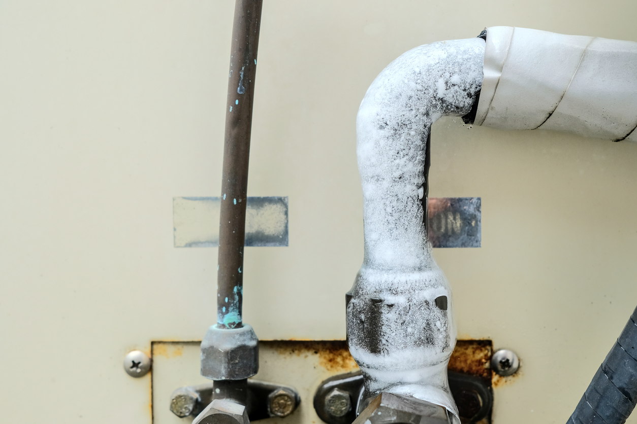 Frost-covered pipe elbow connected to a rusted valve assembly against a beige wall.