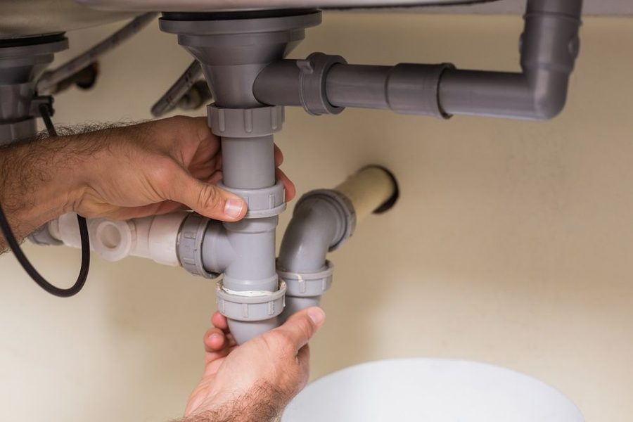 Plumber's hands tightening a gray PVC pipe connection under a sink, working on the P-trap and drainage pipes under the basin.