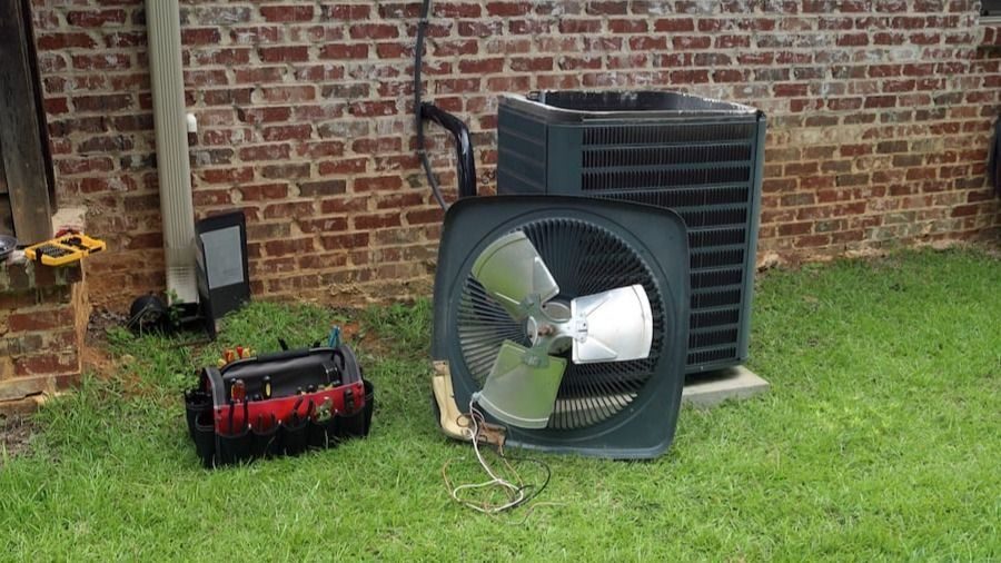 Outdoor air conditioning condenser with a large fan on the lawn beside a brick wall; a red tool bag sits nearby.