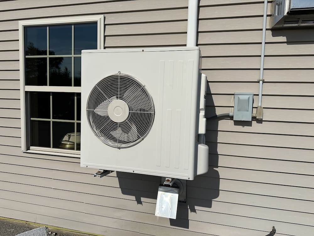 Outdoor HVAC condenser unit mounted on beige siding near a window, with electrical box and conduit nearby