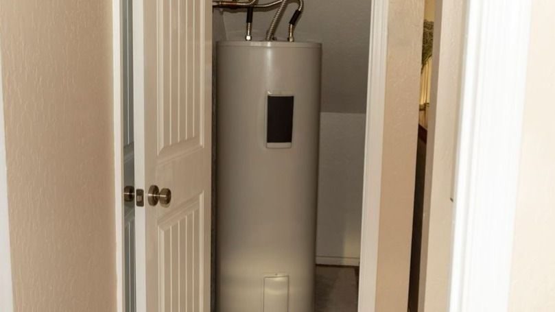 Tall gray water heater in a small utility closet with exposed pipes at the top and a partially open door.